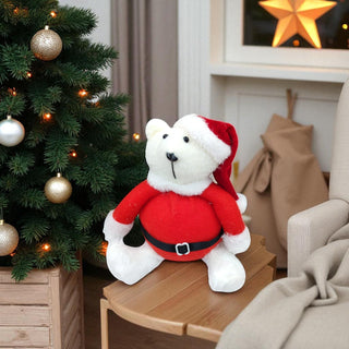 White teddy bear in a Santa outfit on a wooden table with a decorated Christmas tree in the background.