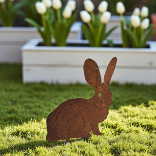 Rust-colored metal rabbit sculpture on grass with a planter of white tulips in the background