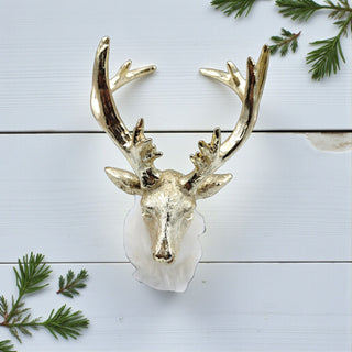Decorative gold deer head mount on a light blue wooden background with greenery.