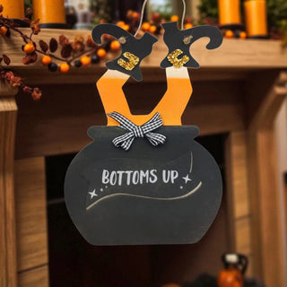 Decorative sign with witches' hats and a pot, featuring the text 'BOTTOMS UP', on a blurred Halloween-themed background.